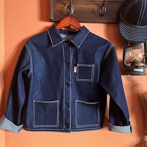 May include: A dark blue denim jacket featuring a classic collar, long sleeves with folded cuffs, and a front button closure. The jacket includes two large square pockets at the waist and one smaller square chest pocket, all accented with white stitching.