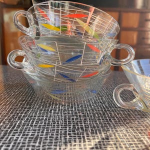 Puede incluir: Una pila de cuatro tazas de té de vidrio transparente con asas. Cada taza presenta un patrón de cuadrícula blanca con acentos coloridos en rojo, amarillo y azul. Las tazas están apiladas sobre una superficie texturizada.