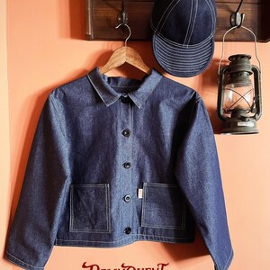 May include: A dark blue denim jacket with a collar, long sleeves, and five black buttons down the front. It features two square front pockets and white contrast stitching. A matching dark blue denim baseball cap with white stitching is also shown.