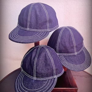 May include: Three dark blue denim caps featuring prominent white contrast stitching along the seams and the curved brim. Each cap has a multi-panel construction with a rounded crown and a short, stiff visor, showcasing a uniform design.