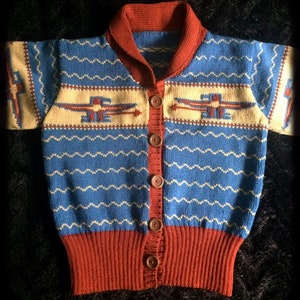 Pode incluir: Cardigã vintage com botões em azul, ferrugem e creme. O suéter apresenta uma bainha e gola caneladas na cor ferrugem, com um corpo azul adornado com padrões de zigue-zague brancos e desenhos geométricos creme. O cardigã tem botões marrons.