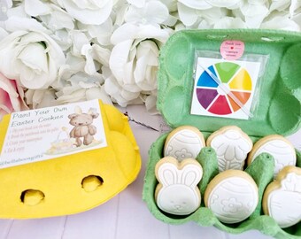 Può includere: Un kit per decorare biscotti a tema pasquale. Il kit comprende sei biscotti a forma di Pasqua, una tavolozza di colori e un pennello. I biscotti sono in un cartone per uova verde e la tavolozza di colori è in un sacchetto di plastica trasparente.
