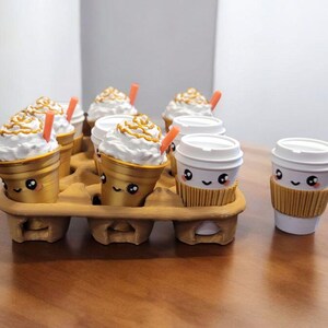 May include: A gold-colored cardboard coffee cup holder with six cups of coffee with whipped cream and a straw. Two additional cups of coffee with whipped cream and a straw are on a wooden table.