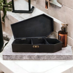 May include: A black coffin-shaped box with a hinged lid and a brass latch. The box is divided into compartments and is sitting on a gray marble surface.