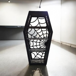 May include: Black coffin-shaped shelf with a spiderweb design. The shelf has four shelves and is made of wood.
