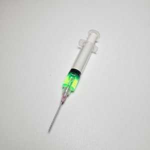 Op de afbeelding: Een doorzichtige plastic spuit gevuld met een levendige groene vloeistof. De spuit heeft een zilveren naald en een witte zuiger. De vloeistof binnenin lijkt een heldere, neon groene kleur te hebben.