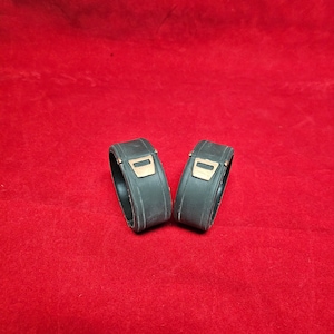 May include: Two black rubber watch bands with gold accents. The bands are designed to fit around a watch case.