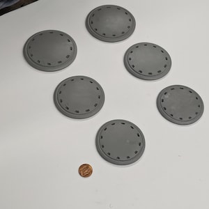 Puede incluir: Seis posavasos circulares grises con un patr&oacute;n decorativo alrededor del borde. Los posavasos est&aacute;n dispuestos en c&iacute;rculo sobre una superficie blanca.