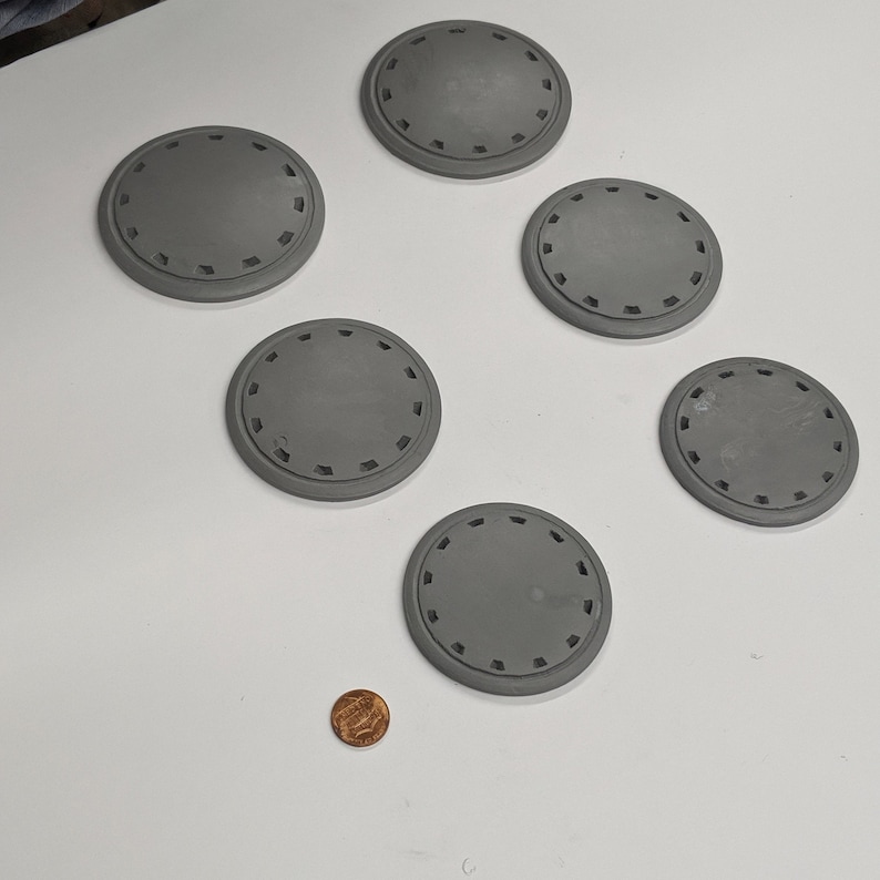 Puede incluir: Seis posavasos circulares grises con un patr&oacute;n decorativo alrededor del borde. Los posavasos est&aacute;n dispuestos en c&iacute;rculo sobre una superficie blanca.