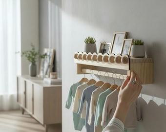 May include: A wooden baby clothes rack with a shelf holding small potted plants and framed photos. Pastel-colored baby clothes hang on wooden hangers. The rack is mounted on a white wall in a well-lit room.