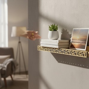 May include: A gold-colored wall shelf with a Greek key design, holding a small potted succulent, several books, and a framed photograph of a beach scene. The shelf is mounted on a neutral-colored wall.