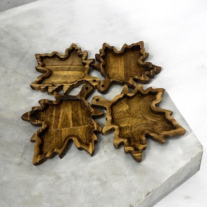 May include: Four wooden leaf-shaped trays with a dark brown finish. The trays are carved with detailed leaf veins and have a shallow, bowl-like interior. The wood grain is visible, adding texture to the decorative pieces.