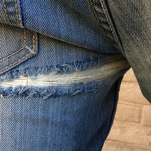 May include: Close-up of a pair of blue denim jeans with a repaired tear. The tear is patched with a white fabric strip.