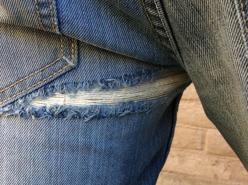 May include: Close-up of a pair of blue denim jeans with a repaired tear. The tear is patched with a white fabric strip.