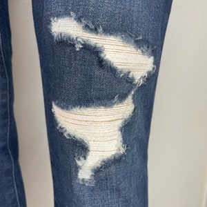 May include: Close-up of a pair of dark blue denim jeans with two distressed areas on the knee. The distressed areas are frayed and have a white fabric underneath.
