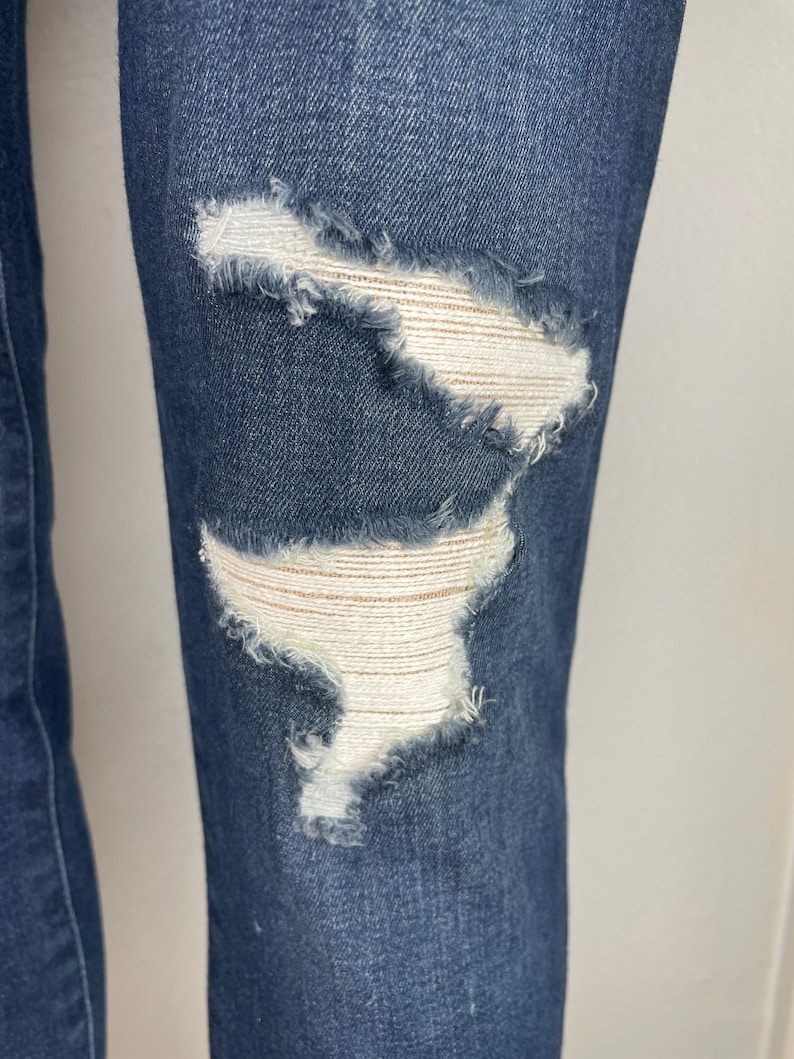 May include: Close-up of a pair of dark blue denim jeans with two distressed areas on the knee. The distressed areas are frayed and have a white fabric underneath.