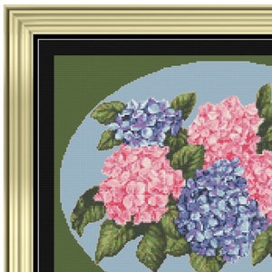 Puede incluir: Obra de arte de punto de cruz de hortensias rosas y azules con hojas verdes, sobre un óvalo azul claro sobre un fondo verde. La pieza enmarcada tiene un marco dorado.