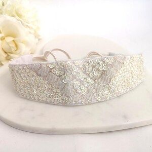 May include: A white beaded headband with a sequined design. The headband has a scalloped pattern and is decorated with small, round sequins. The elastic band is white. The headband is suitable for a special event.