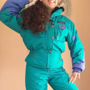 Puede incluir: Un traje de esqu&iacute; turquesa con ribetes morados y azules. El traje tiene una cremallera en la parte delantera y una capucha.