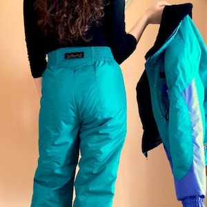 Puede incluir: Pantalón de esquí de color turquesa con cintura negra y cremallera lateral. El pantalón está hecho de un material brillante y resistente al agua.