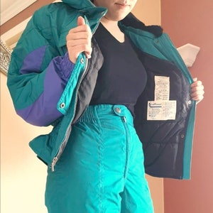 Puede incluir: Un traje de esqu&iacute; azul turquesa con una chaqueta morada y negra. La chaqueta tiene una cremallera y una capucha. El pantal&oacute;n tiene cintura alta y pierna recta.