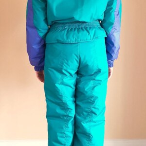 Puede incluir: Un traje de esqu&iacute; turquesa con detalles morados. El traje tiene una cremallera en la parte delantera y un cintur&oacute;n en la cintura.