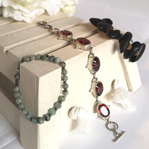 Può includere: Tre braccialetti su un espositore in legno bianco. Un braccialetto è fatto di perline verdi e nere, un altro è fatto di argento con pietre preziose rosse e il terzo è un braccialetto di perline nere con un accento in legno.