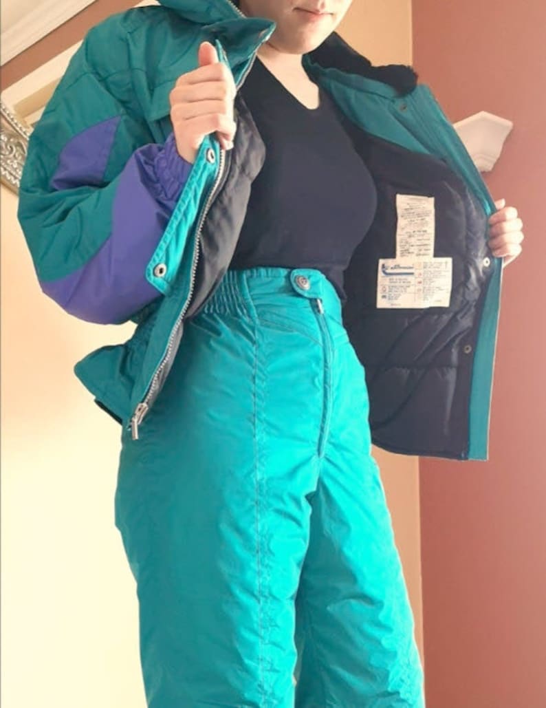 Puede incluir: Un traje de esqu&iacute; azul turquesa con una chaqueta morada y negra. La chaqueta tiene una cremallera y una capucha. El pantal&oacute;n tiene cintura alta y pierna recta.