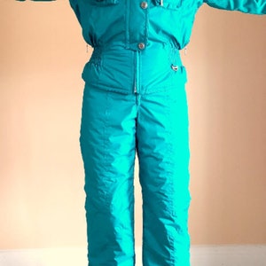 Puede incluir: Un traje de esqu&iacute; turquesa con detalles azules y una cremallera en la parte delantera. El traje tiene una capucha y un bolsillo en el pecho izquierdo.