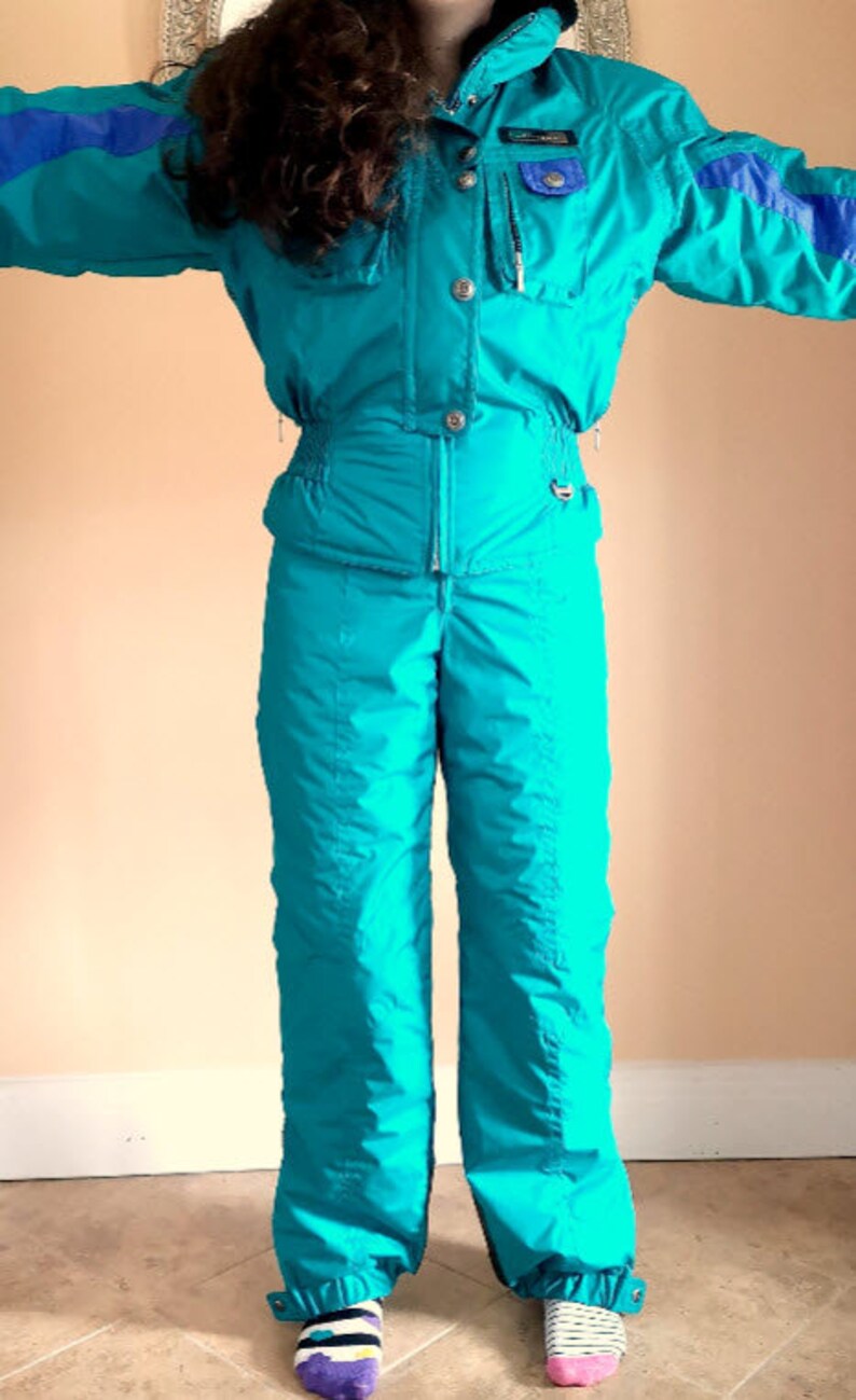 Puede incluir: Un traje de esqu&iacute; turquesa con detalles azules y una cremallera en la parte delantera. El traje tiene una capucha y un bolsillo en el pecho izquierdo.
