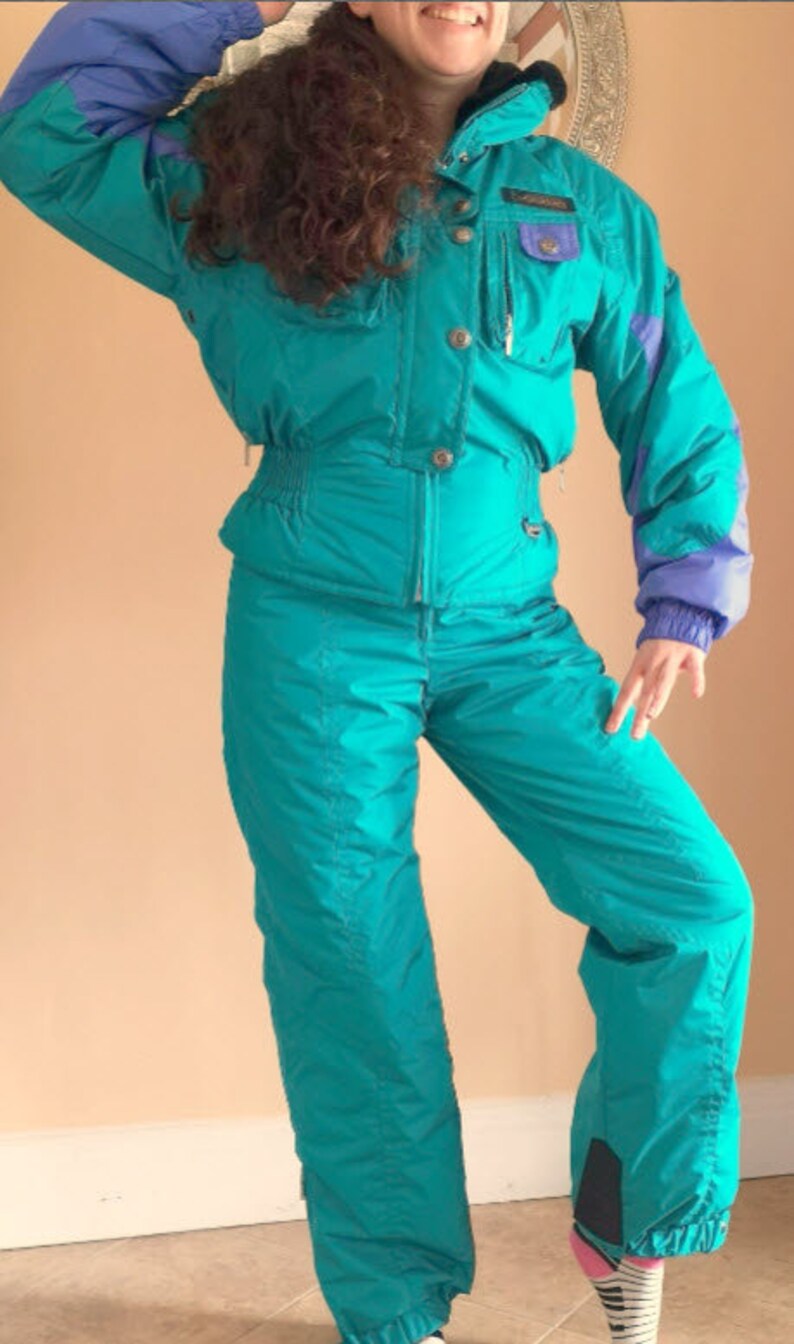 Puede incluir: Un traje de esqu&iacute; turquesa con ribetes morados y azules. El traje tiene una cremallera en la parte delantera y una capucha.