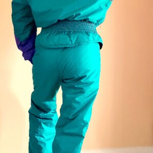 Puede incluir: Un traje de esqu&iacute; turquesa con cuello negro y detalles azules. El traje tiene una cremallera en la parte delantera y una cintura ce&ntilde;ida.