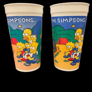 May include: Two tall plastic cups with white rims and a colorful design featuring The Simpsons family camping. The cups have a blue background with the words "SIMPSONS" and "THE SIMPSONS" printed on them.