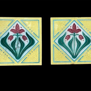 May include: Two square ceramic tiles featuring a central diamond motif with a stylized maroon flower and dark green leaves. The background is a pale yellow with a visible crackle glaze finish, framed by thin teal lines and small dots.