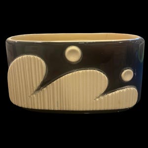 May include: An oval ceramic planter with a glossy dark brown finish. The exterior features raised, cream-colored textured panels with vertical ribbing and two small circular accents, contrasting against the smooth, dark brown glazed surface.
