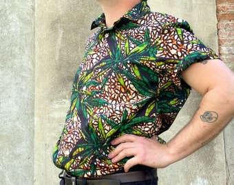 May include: Short-sleeved button-down shirt with a vibrant green and brown leaf pattern on a black background. The shirt has a classic collar and is paired with a black belt and dark wash jeans. The overall style is casual and eye-catching.