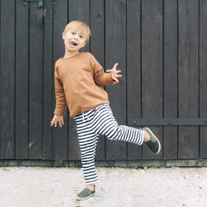 May include: A young child wearing a brown long-sleeved shirt and black and white striped pants is standing in front of a wooden door. The child is smiling and has their leg raised.