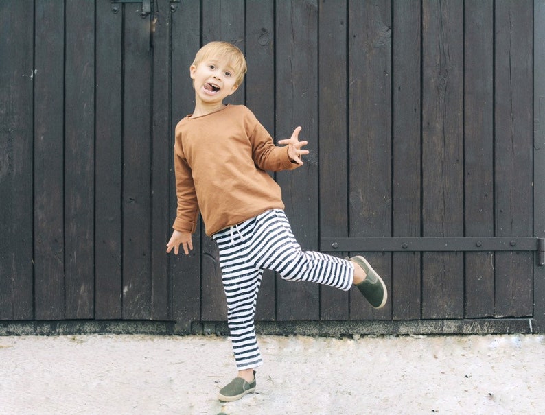 May include: A young child wearing a brown long-sleeved shirt and black and white striped pants is standing in front of a wooden door. The child is smiling and has their leg raised.