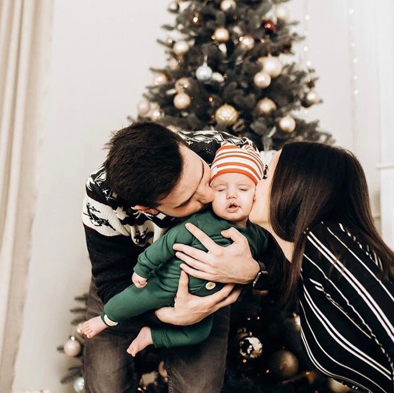 May include: A baby wearing a solid green long-sleeved onesie with a single button and a red and white striped beanie, held by two adults, one in a black and white patterned jumper and the other in a black and white striped long-sleeved top.