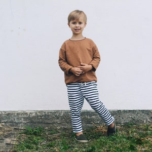 May include: A young boy wearing a brown long-sleeved shirt and black and white striped pants stands in front of a white wall. He is smiling at the camera.