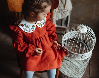Op de afbeelding: Een jong meisje in een rode jurk met een witte kraag versierd met kleurrijke bloemen en vogels zit op een houten stoel. Ze draagt ​​blauwe panty's en witte schoenen. Een witte vogelkooi staat naast de stoel.