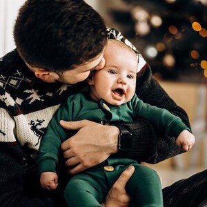 May include: A long-sleeved dark green one-piece outfit for an infant, featuring a round neckline, ribbed cuffs at the wrists and ankles, and two light-coloured wooden buttons centred on the front placket, providing a simple and functional design.
