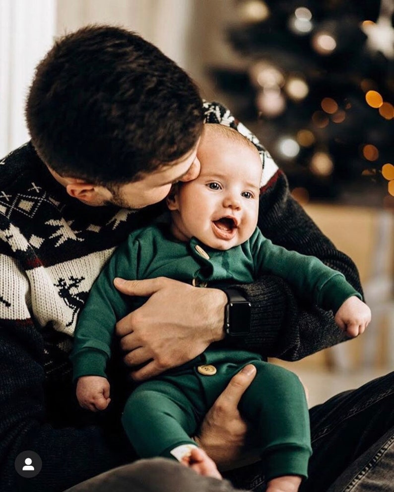 May include: A long-sleeved dark green one-piece outfit for an infant, featuring a round neckline, ribbed cuffs at the wrists and ankles, and two light-coloured wooden buttons centred on the front placket, providing a simple and functional design.