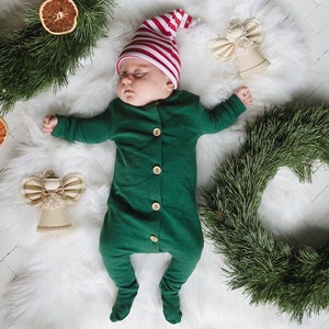 May include: A sleeping infant wearing a long-sleeved, footed forest green onesie with four light-coloured wooden buttons down the front and a red and white striped knit hat with a white pom-pom, surrounded by pine branches, dried orange slices, and two fabric angels.