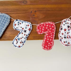 May include: A fabric banner featuring the numbers 1, 7, 7, and 6, each cut into a numeral shape with scalloped edges. The numbers are strung on a white cord, displaying patterns of blue with white crosses, white with red and blue stars, and red with white flowers.