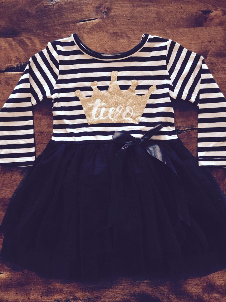 May include: A black and white striped long-sleeved dress with a black tulle skirt. The dress has a gold glitter crown with the word "two" on it.