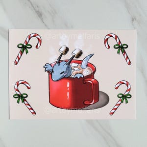 Puede incluir: Una ilustración caprichosa presenta una criatura azul relajándose en una taza roja de chocolate caliente con malvaviscos. Bastones de caramelo con lazos verdes rodean la taza. El texto "@artbymelfaris" es visible.