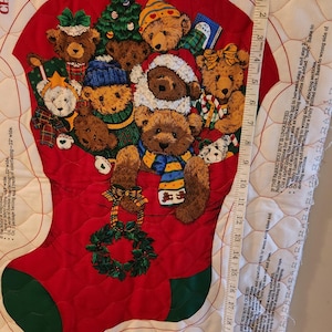 May include: A red fabric Christmas stocking panel featuring a group of brown teddy bears, a green wreath, and a Christmas tree. A white measuring tape is placed vertically along the right side, showing a length of approximately 22 inches.