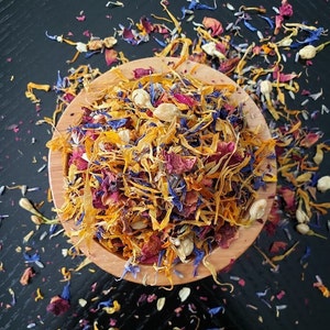 May include: A wooden bowl filled with dried flower petals in shades of yellow, orange, red, purple, and blue. The petals are scattered around the bowl on a black surface.