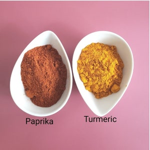 May include: Two small white bowls, one filled with red paprika powder and the other with yellow turmeric powder. The bowls are on a pink background. The words "Paprika" and "Turmeric" are written below the bowls.